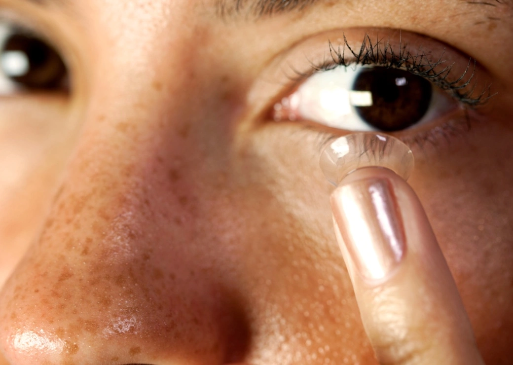 A close up of a Caucasian female holding a translucent contact lens on her index finger, in front of her eye, moments before application.